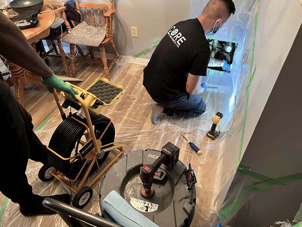 Core Plumbing employees at work: One kneeling and cleaning a kitchen stack, while the other uses a yellow camera tool to inspect the pipe's cleanliness.